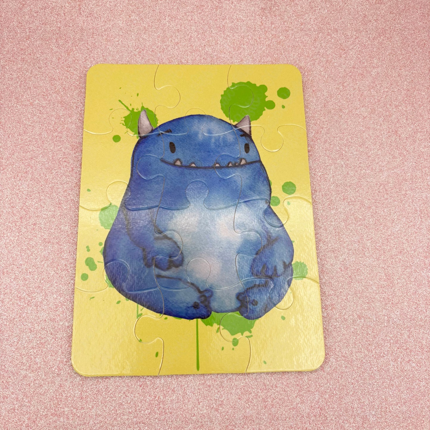 Jigsaw puzzle with a blue monster design on a yellow background -jigsaw is laying against a pink surface