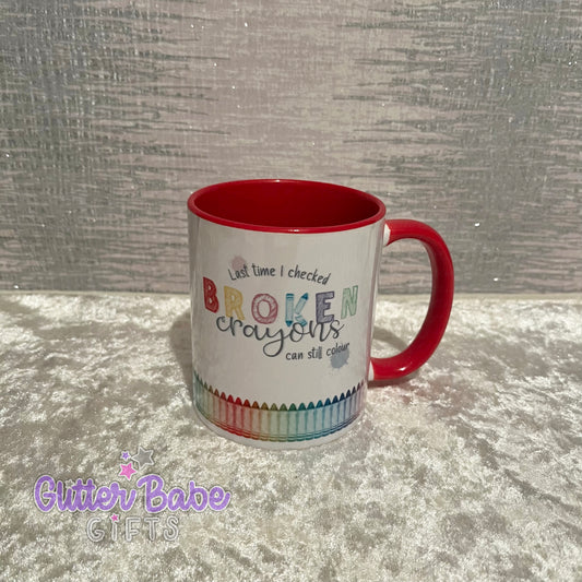 A white ceramic mug with a red handle and inner part. The mug has colourful text and designs resembling a broken crayon pattern around the bottom.