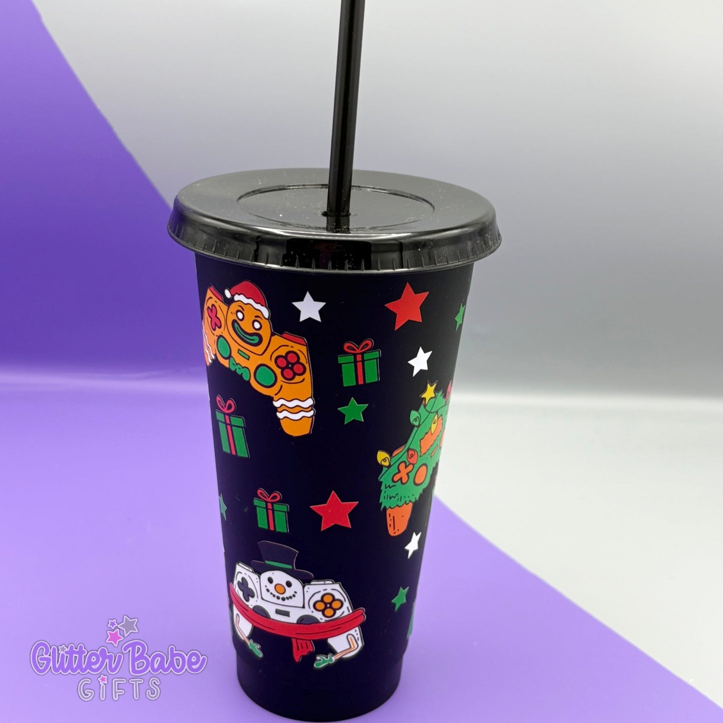 A Black cold cup against a grey and purple backdrop. Cold Cup design has gaming controllers designed to look like snowmen, Christmas trees and gingerbread men on it alongside parcels and stars.