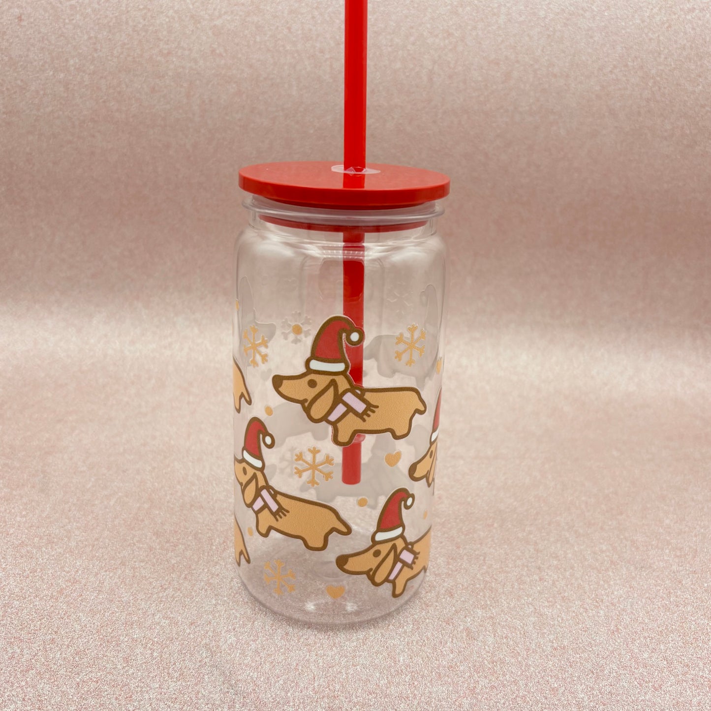 Clear tumbler with red lid and straw featuring Christmas-themed dachshund illustrations on a light pink background.