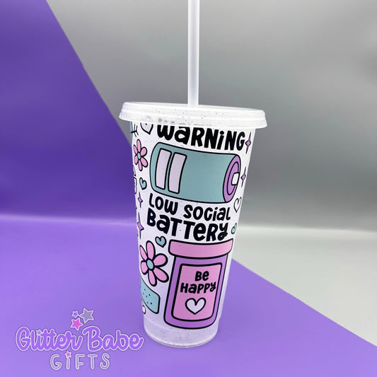 A frosted glitter cold cup showing a warning low social battery image with hearts and flowers, Cup is on a purple and grey background.