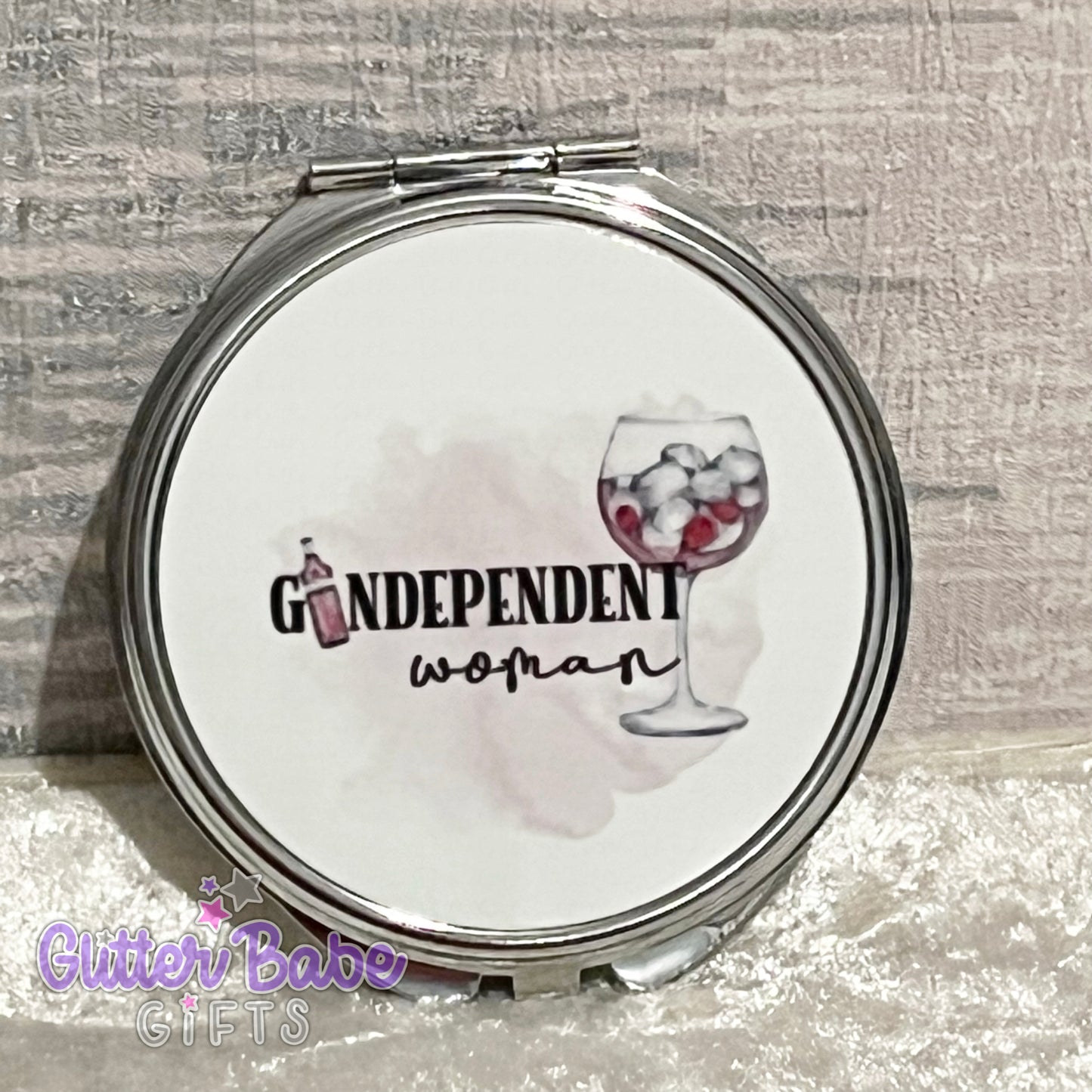 A handheld compact mirror with a silver rim and a design featuring the text 'Gindependant Woman' along with an illustration of a glass of gin and ice.