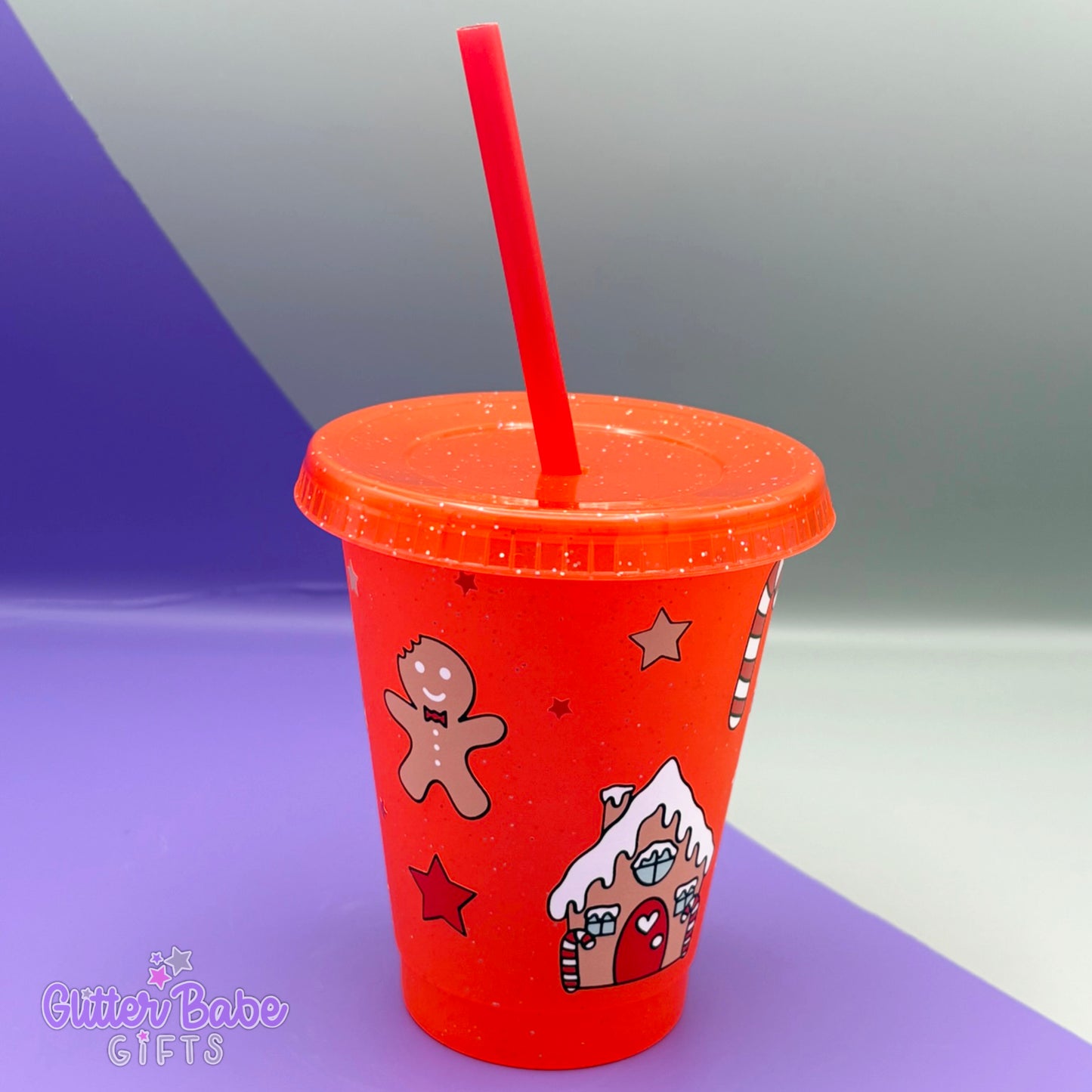 A mini red glitter cold cup with gingerbread men, stars and gingerbread house design on it. The cup sits in front of a purple and grey background.