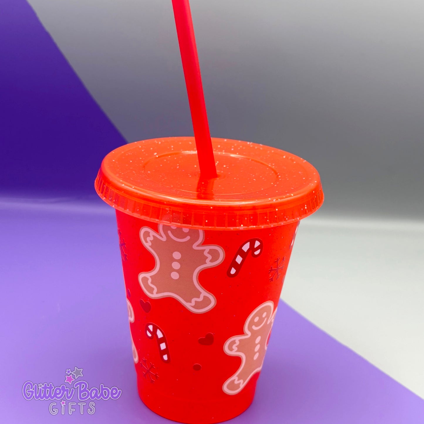 A mini red glitter cold cup with gingerbread men, hearts and candycanes design on it. The cup sits in front of a purple and grey background.