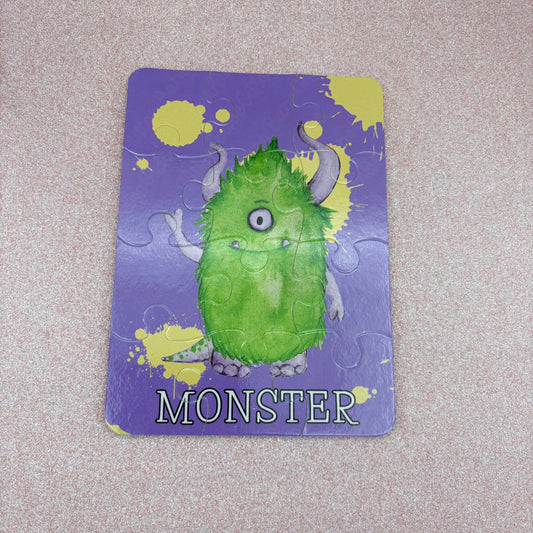 Jigsaw puzzle with a green monster illustration on a purple background laying against a pink surface