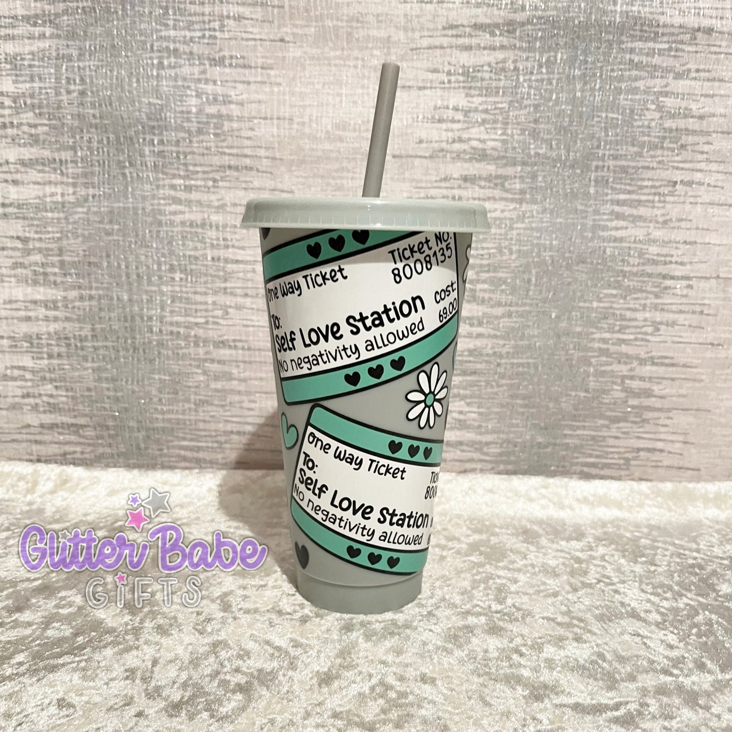 A grey cold cup with a travel ticket style pattern and the text 'Self Love Station' printed on it.