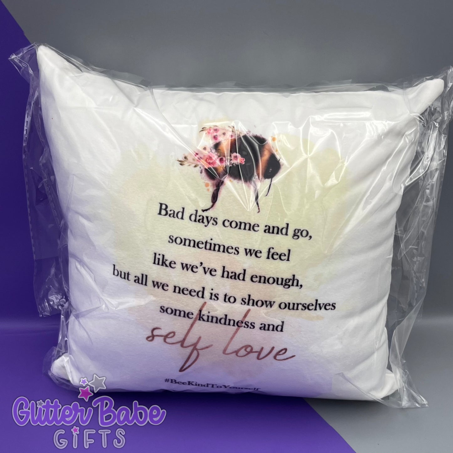 A cushion in cellophane bag showing a bee design and text. cushion is against a purple and grey background