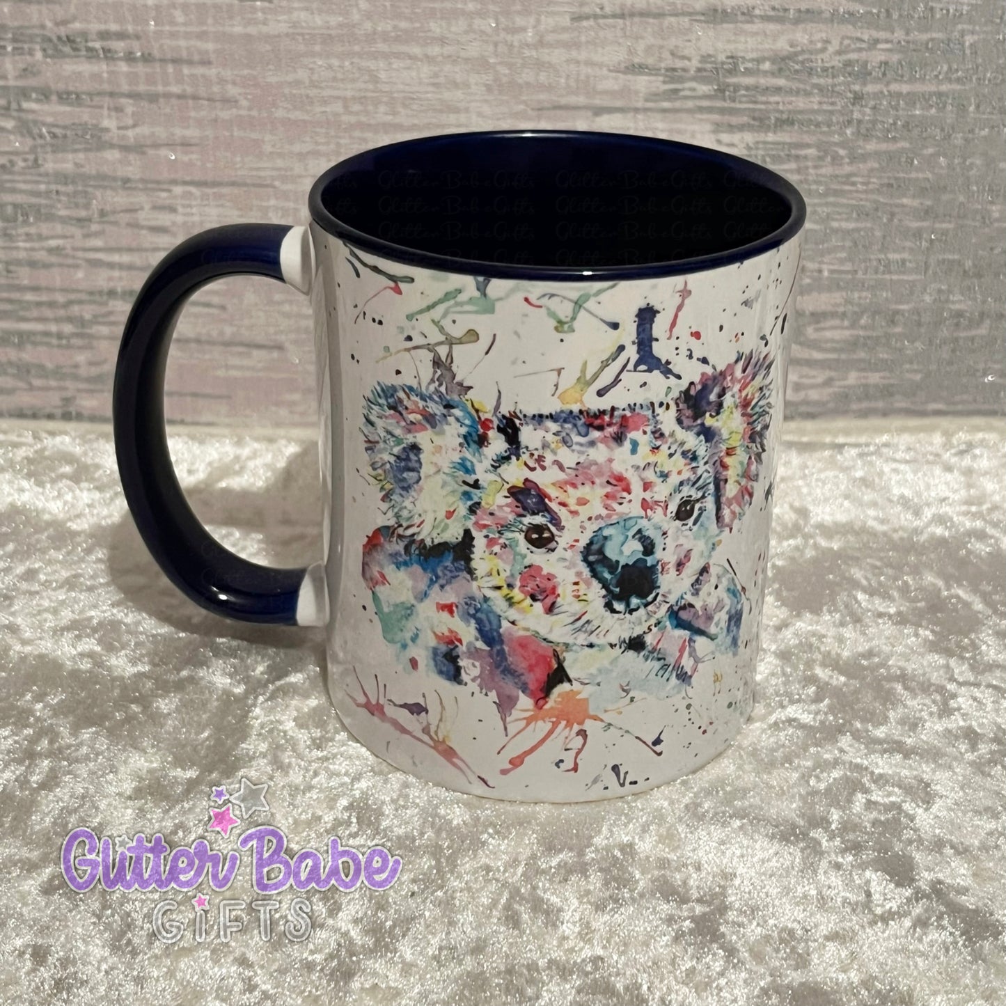 A ceramic mug with a multicolored koala design on the front, with a dark blue inner surface and handle, displayed on a textured background.