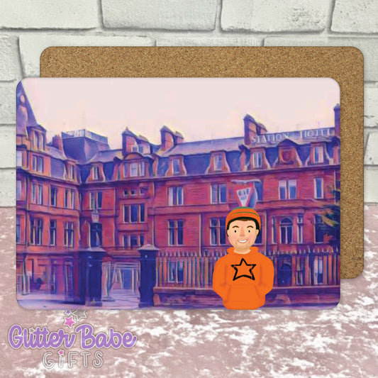 A square placemat with an illustration of the Ayr Station Hotel, and a cartoon character wearing an orange shirt with a star.