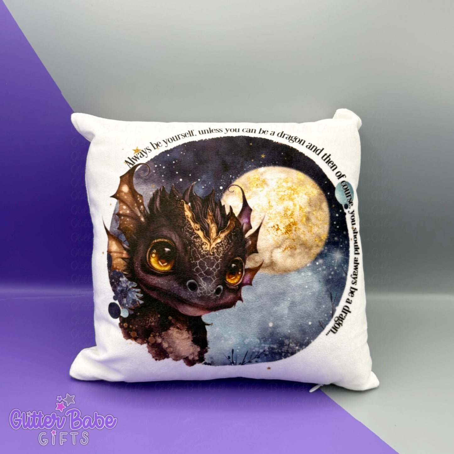 A white cushion with a black and gold dragon against a night sky and moon with wording "always be yourself unless you can be a dragon and then of course you should always be a dragon "printed on it. The cushion sits against a purple and grey background.