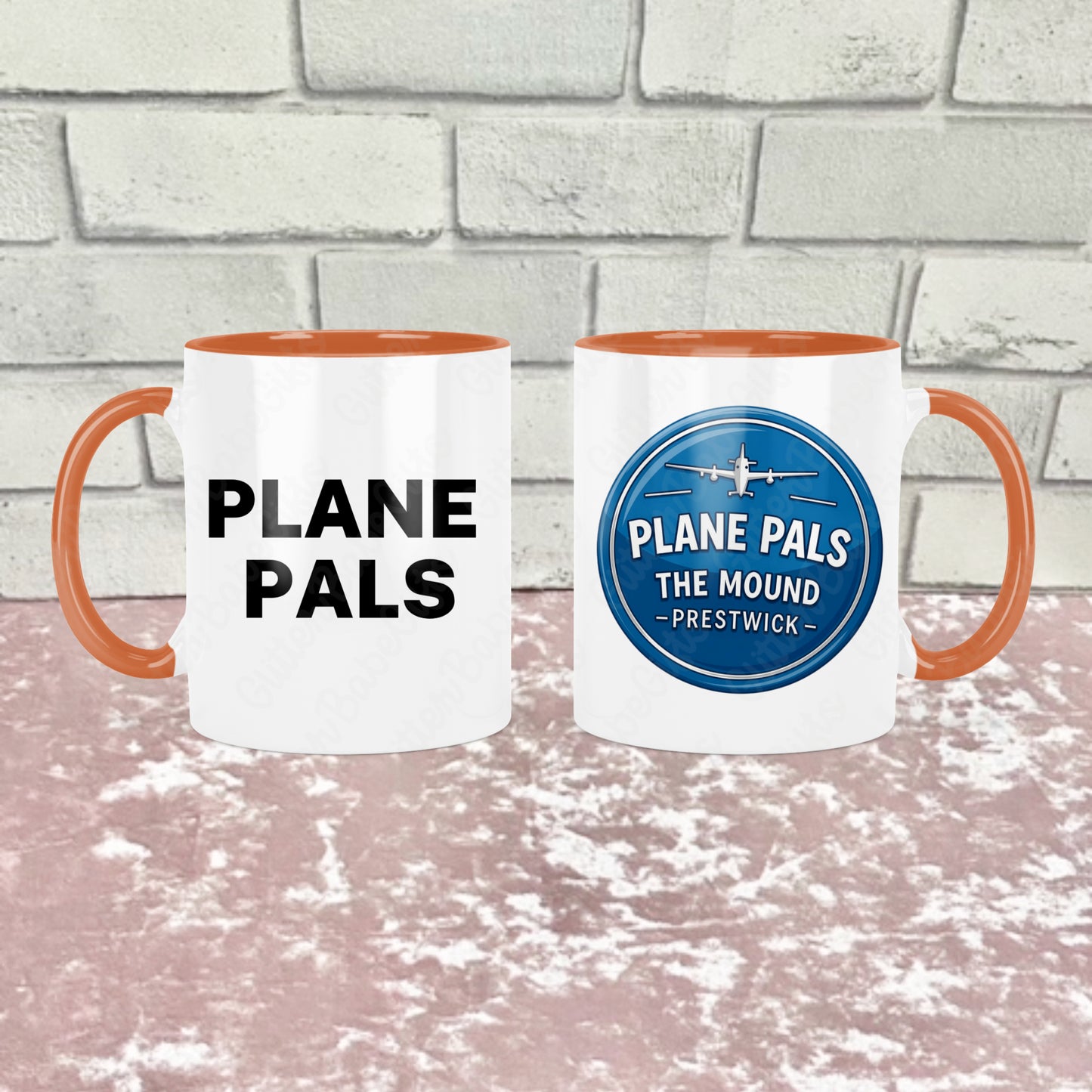 Two white ceramic mugs with orange handles and orange interiors show side by side with one side reading 'Plane pals' in black text and the other reading 'PLANE PALS THE MOUND - PRESTWICK' and an airplane graphic. They are shown side by side to show both sides of the mug, the background is a pink textured surface in front of a light gray brick wall.