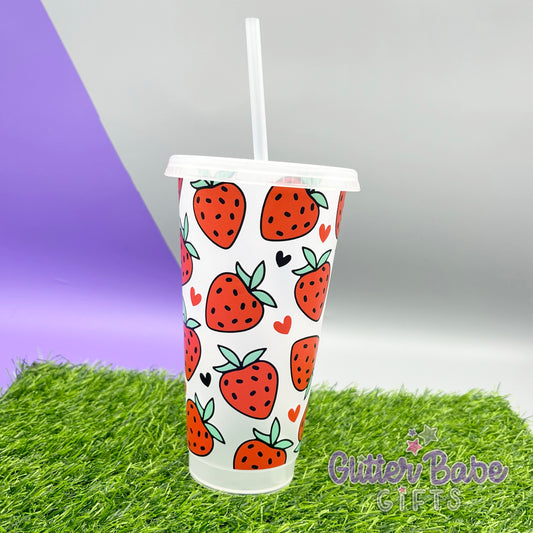 A clear plastic cold cup with a textured strawberry design in red set against a purple background with a grassy surface.