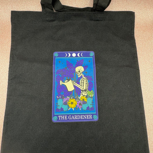 Black tote bag with 'The Gardener' tarot card design featuring a skeleton watering plants.