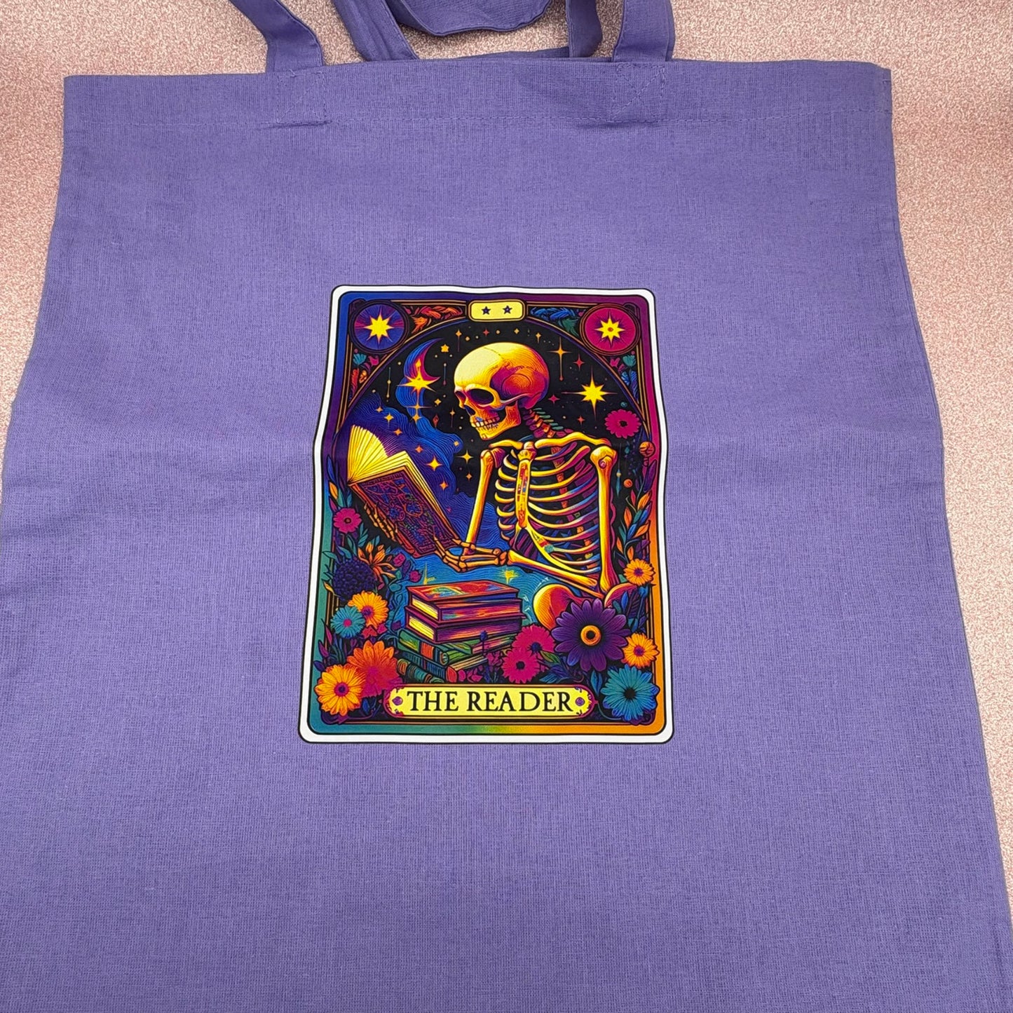 Purple tote bag with a colorful skeleton design labeled 'The Reader' on a pink surface.