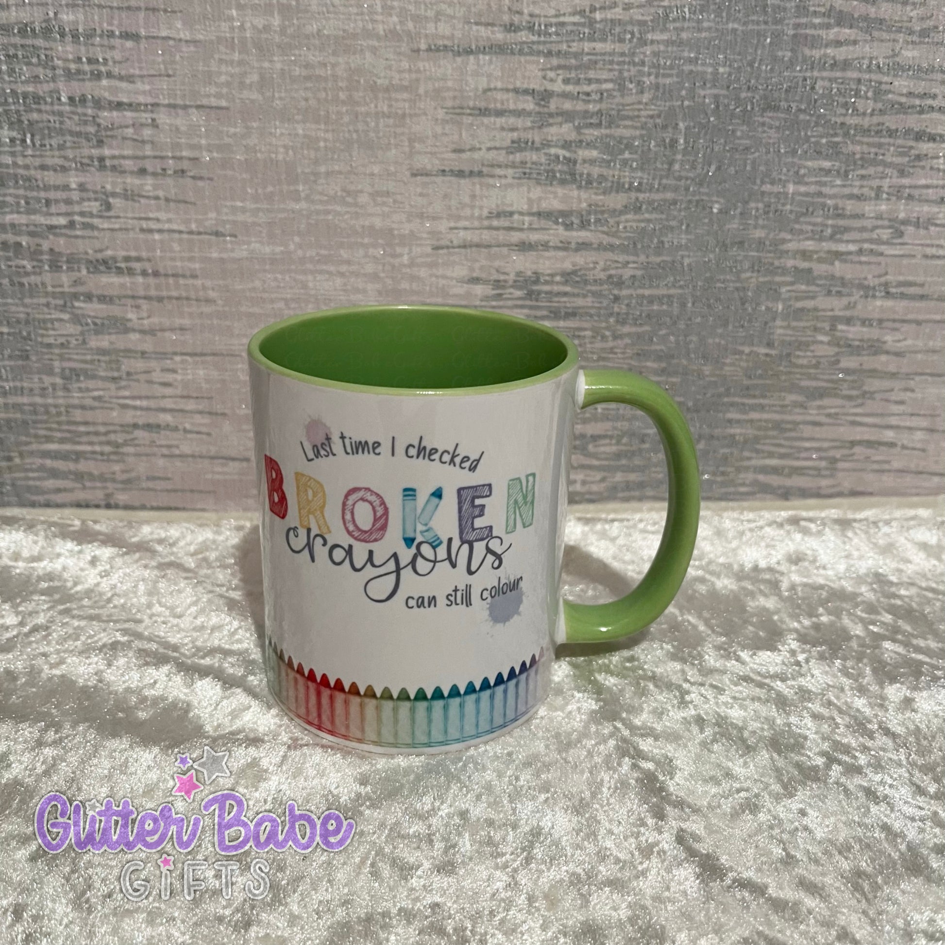 A white ceramic mug with a green handle and inner part. The mug has colourful text and designs resembling a broken crayon pattern around the bottom.