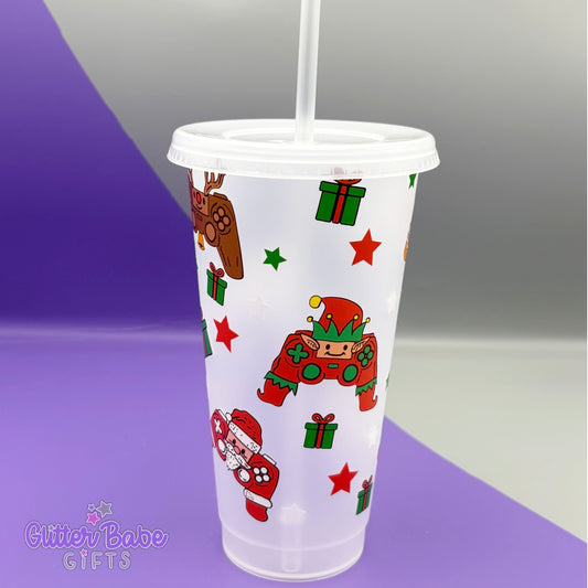 A Frosted Clear  cold cup against a grey and purple backdrop. Cold Cup design has gaming controllers designed to look like snowmen, Christmas trees and gingerbread men on it alongside parcels and stars.