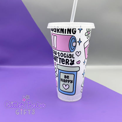 A frosted glitter cold cup showing a warning low social battery image with hearts and stars, Cup is on a  purple and grey background. 