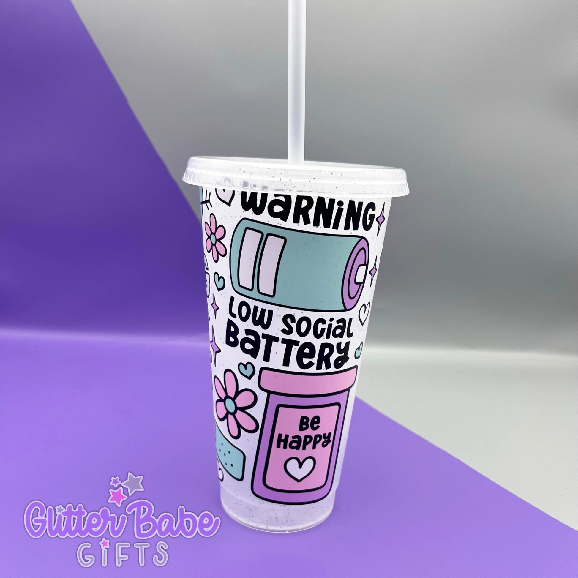 A frosted glitter cold cup showing a warning low social battery image with hearts and flowers, Cup is on a  purple and grey background. 