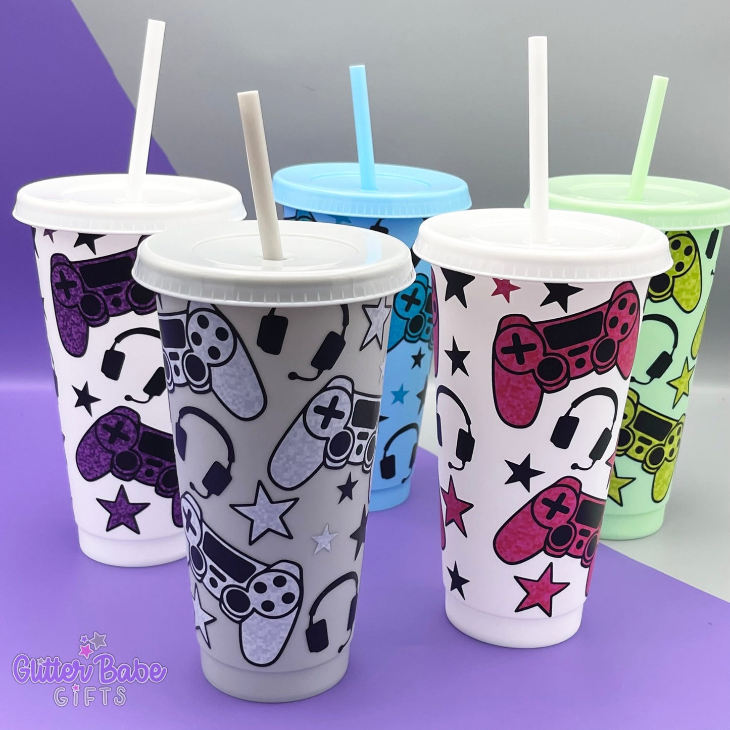 Five cold cups of varying colours with gaming controller design on them against a purple and grey backdrop