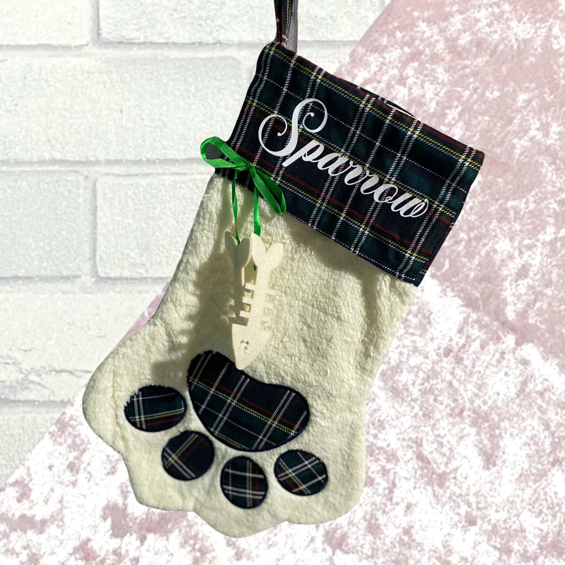 Green tartan cuffed white fleecy stocking with tartan paw print design and a green ribbon with fish bone shape hanging from it with name in white on tartan cuff