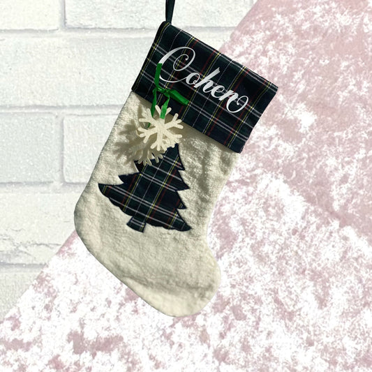 white fleecy boot shaped stocking with green tartan trim with name in white writing. Stocking body has a green tartan tree applique. a green ribbon and white snowflake hangs from the tartan sock trim. Stocking is against a white and pink backdrop