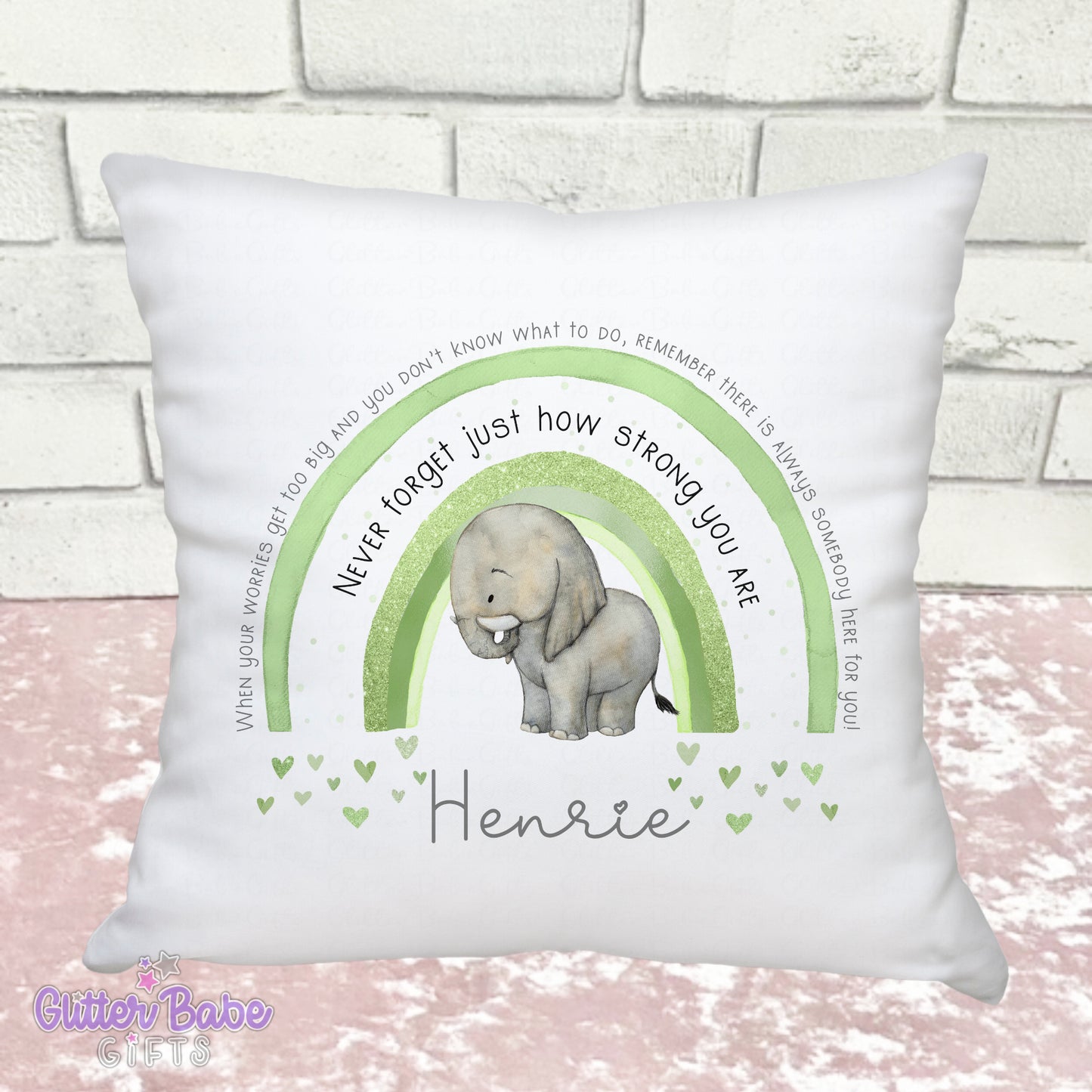 white cushion with a green rainbow and an elephant with writing on it in front of a pink and white brick background.