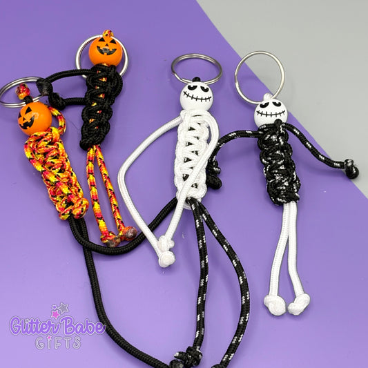 A selection of paracord People keyrings. Mixture of white black and orange paracords used with white or orange wooden pumpkin inspired beads for heads. these a laying against a purple and grey background
