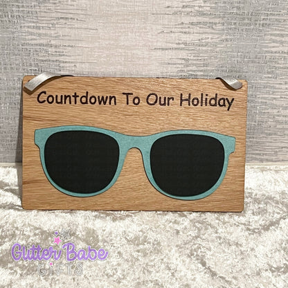 A wooden chalkboard sign with a 'Countdown to our Holiday' message, designed to look like sunglasses, placed on a textured surface.