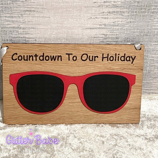 A wooden sign with painted sunglasses chalkboard saying 'countdown to our holiday'. Board is against a textured backdrop