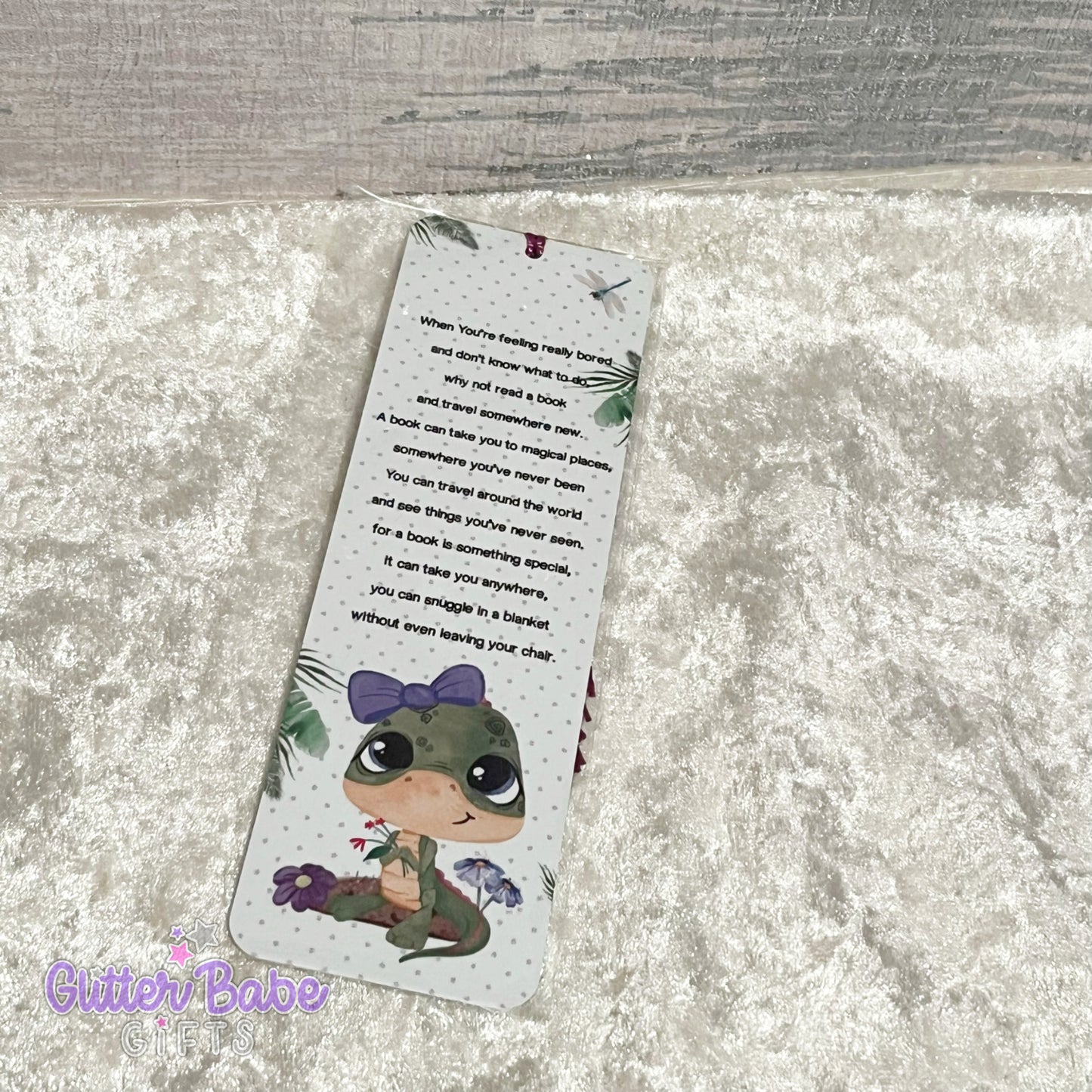A metal bookmark with a cute dinosaur wearing a bow illustration and a floral pattern. The bookmark has tassels on the top edge, colour of which is random.