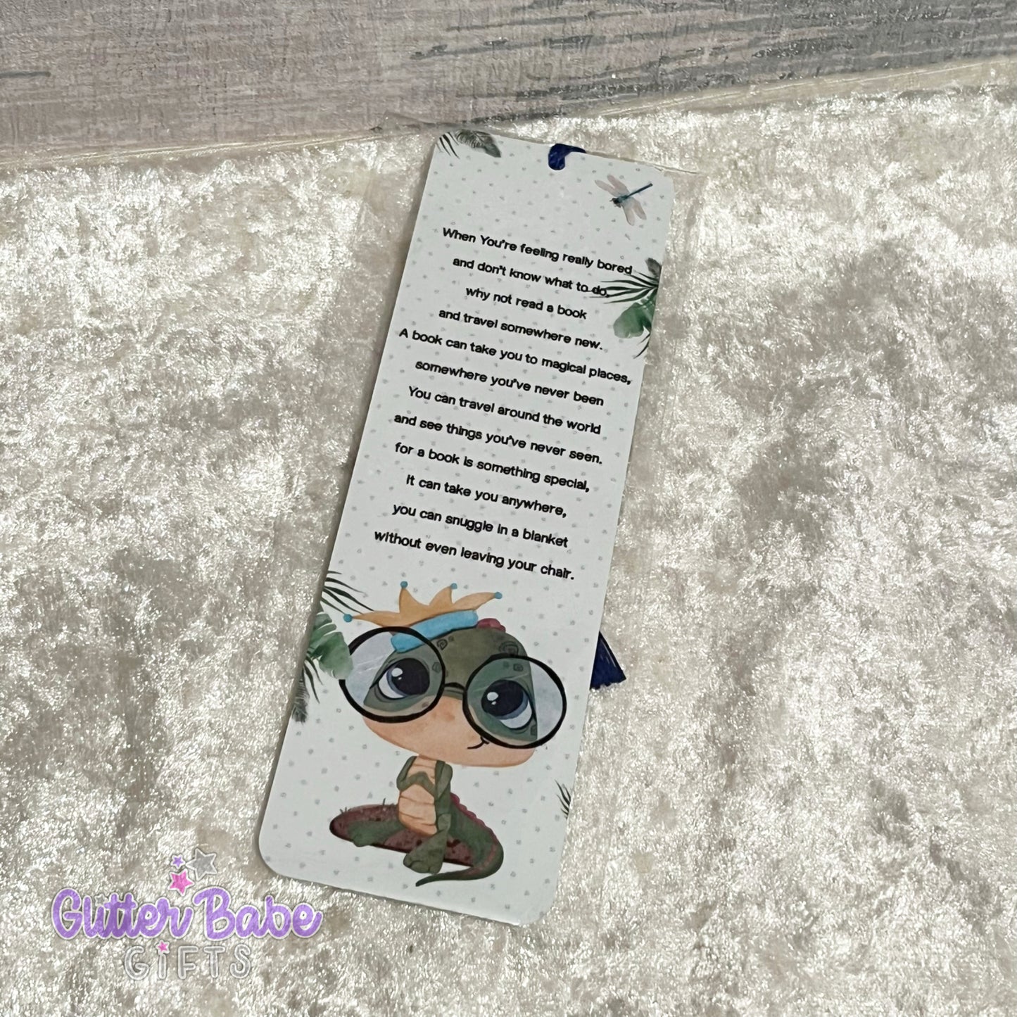A metal bookmark with a cute dinosaur wearing glasses illustration and a leafy pattern. The bookmark has tassels on the top edge, colour of which is random.