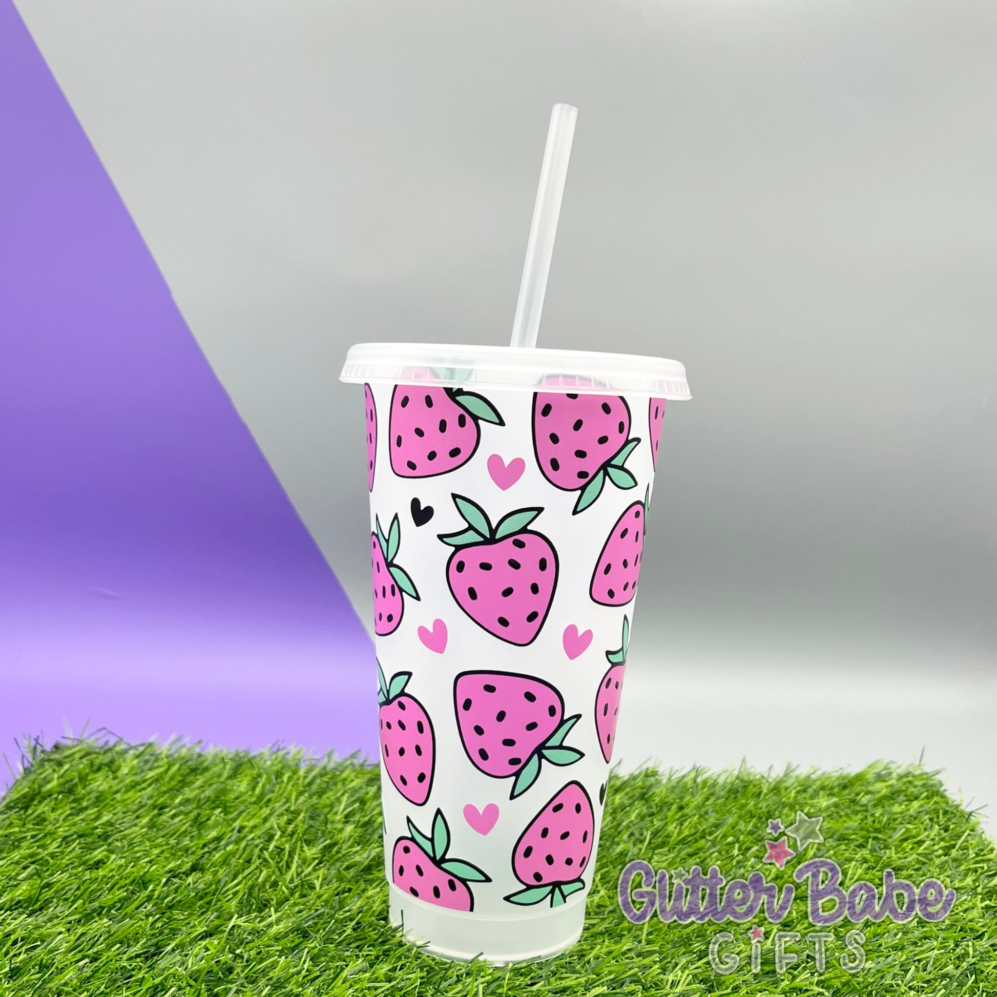 A clear plastic cold cup with a textured strawberry design in pink,  set against a purple background with a grassy surface.