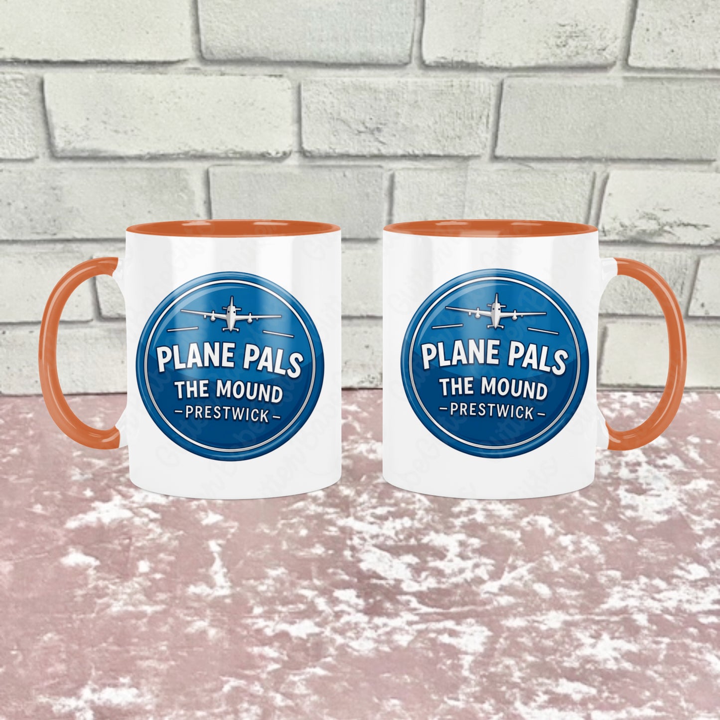 Two white ceramic mugs with orange handles and orange interiors, each printed with a blue circular logo reading 'PLANE PALS THE MOUND - PRESTWICK' and an airplane graphic. They are shown side by side to show how the mug  looks on both sides the background is a pink textured surface in front of a light gray brick wall. 