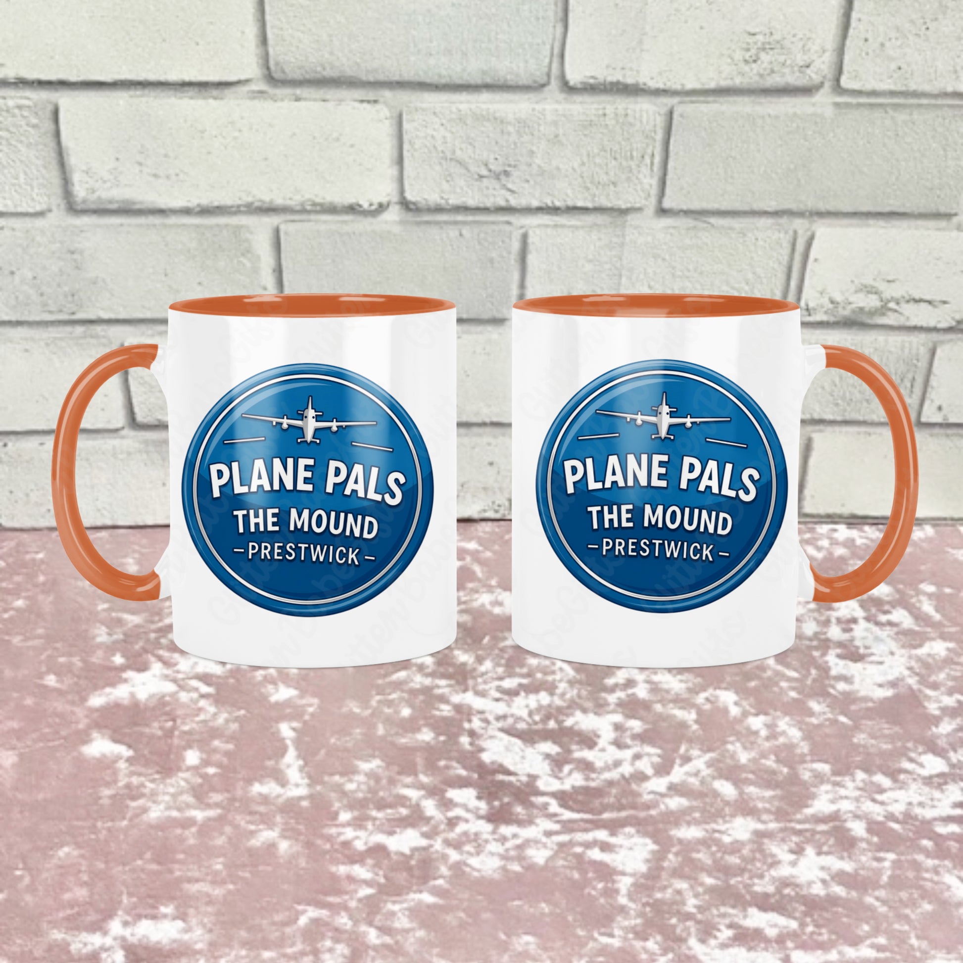 Two white ceramic mugs with orange handles and orange interiors, each printed with a blue circular logo reading 'PLANE PALS THE MOUND - PRESTWICK' and an airplane graphic. They are shown side by side to show how the mug  looks on both sides the background is a pink textured surface in front of a light gray brick wall. 