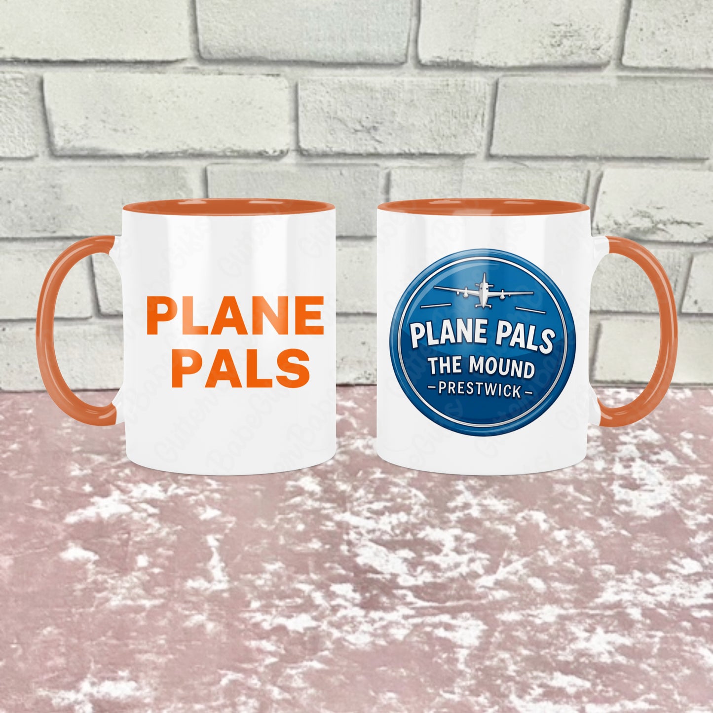 Two white ceramic mugs with orange handles and orange interiors show side by side with one side reading 'Plane pals' in orange text and the other reading 'PLANE PALS THE MOUND - PRESTWICK' and an airplane graphic. They are shown side by side to show both sides of the mug, the background is a pink textured surface in front of a light gray brick wall. 