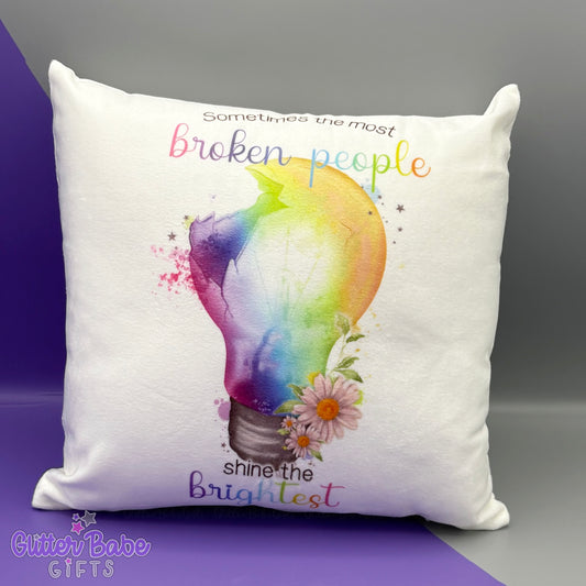 a white cushion with a rainbow coloured lightbulb design with flowers and text against a purple and grey backdrop. 