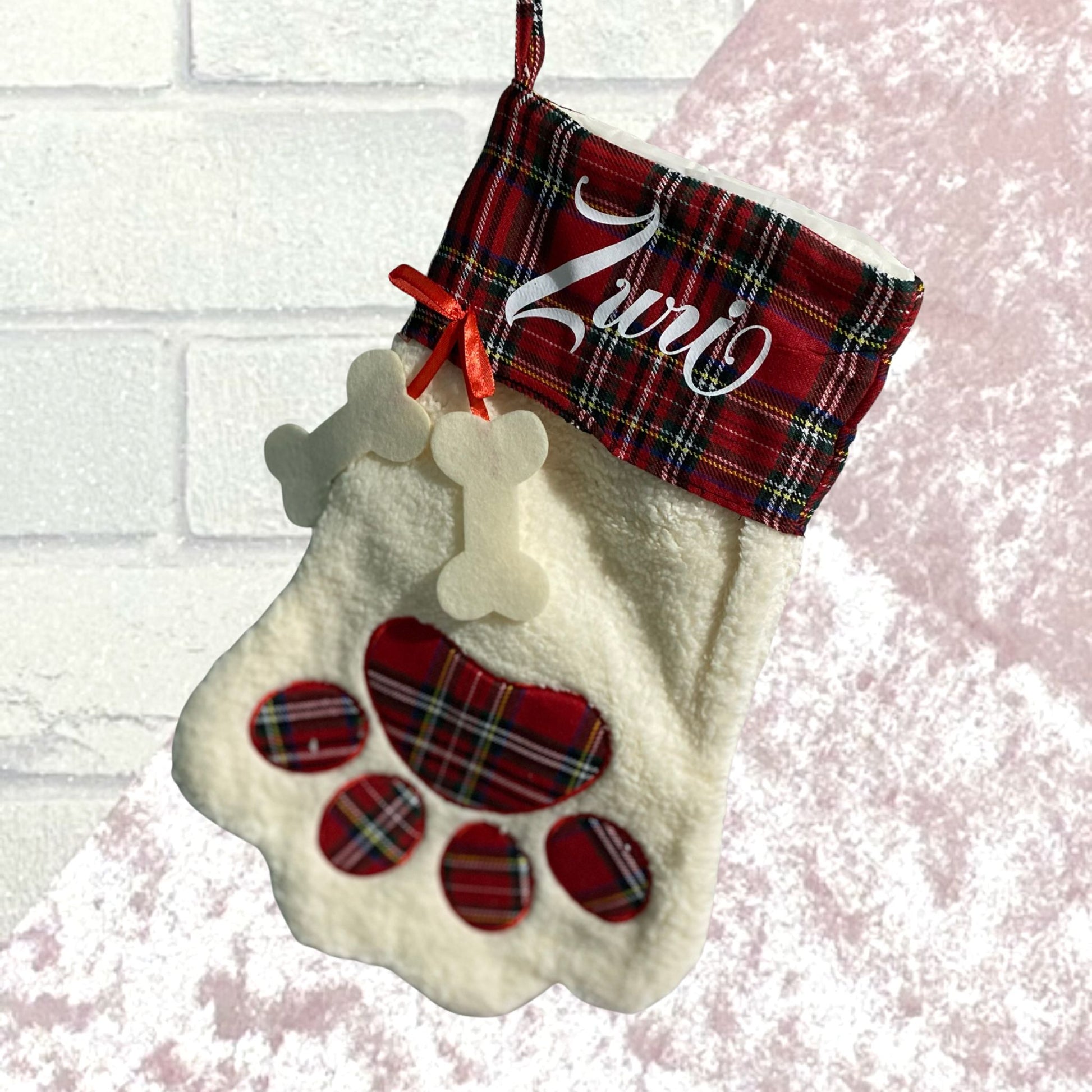 Red tartan cuffed white fleecy stocking with tartan paw print design and a red ribbon with  bone shape hanging from it with name in white on tartan cuff