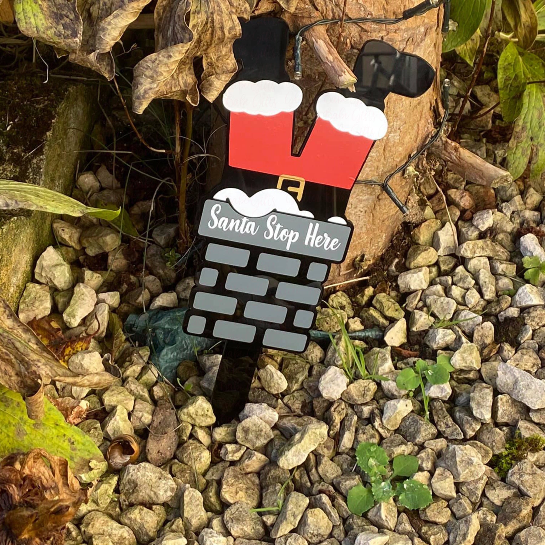 Black acrylic sign in the garden in the shape of santa's legs hanging out a grey brick chimney with white writing